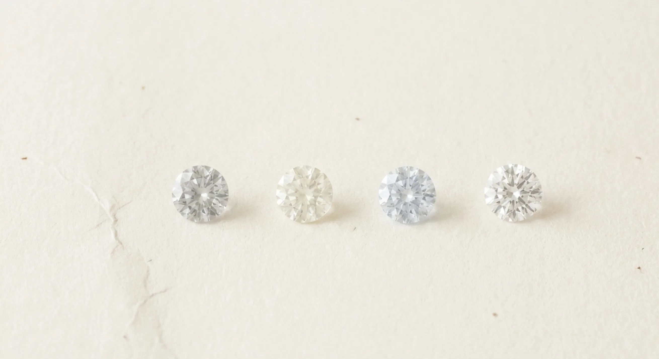 Four loose white sapphires on white paper showing faint grey, yellow and blue tints beside one truly colorless stone
