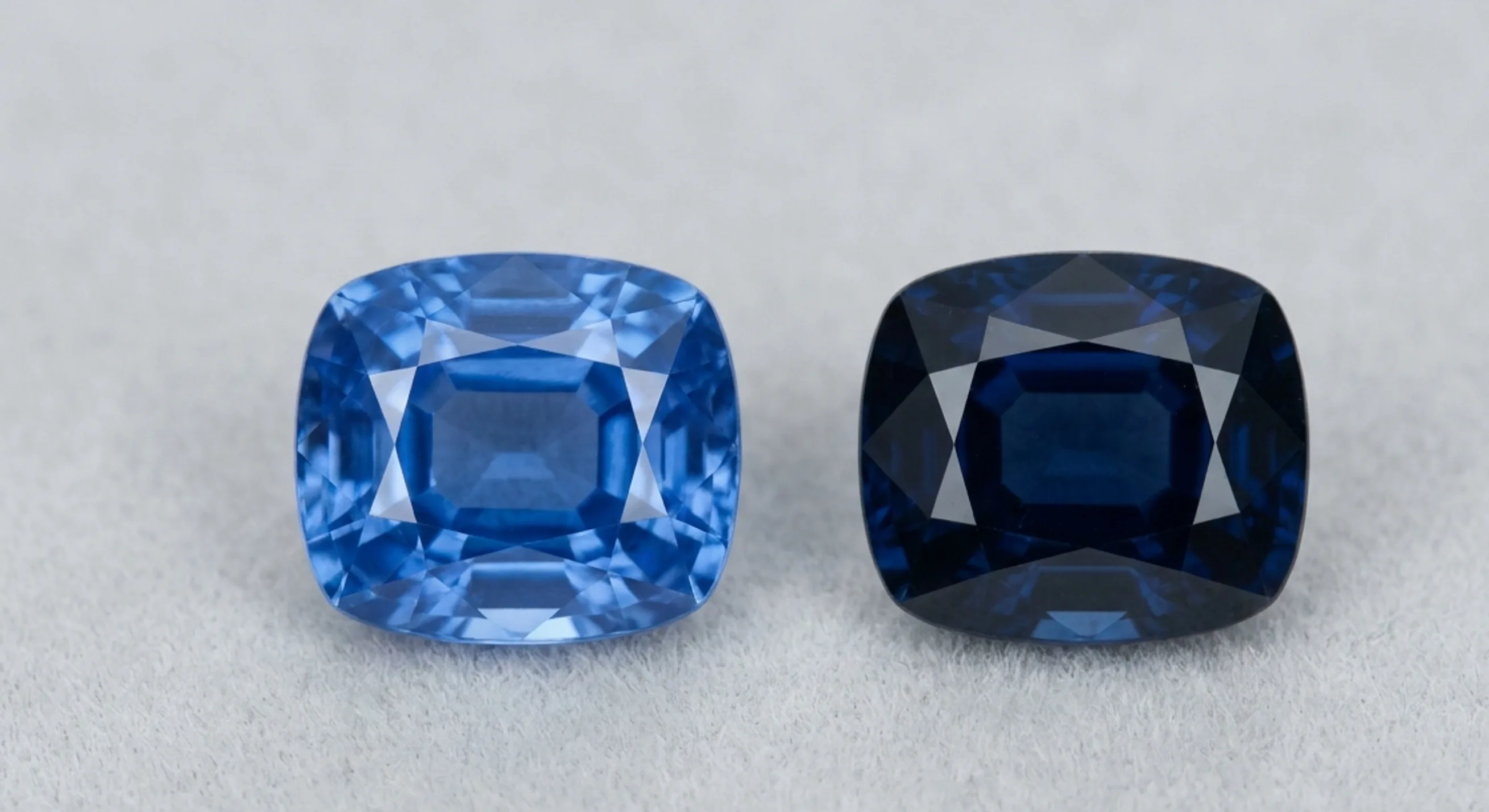 Two loose blue sapphires side by side, one vivid medium-tone and one darker over-saturated, under identical lighting