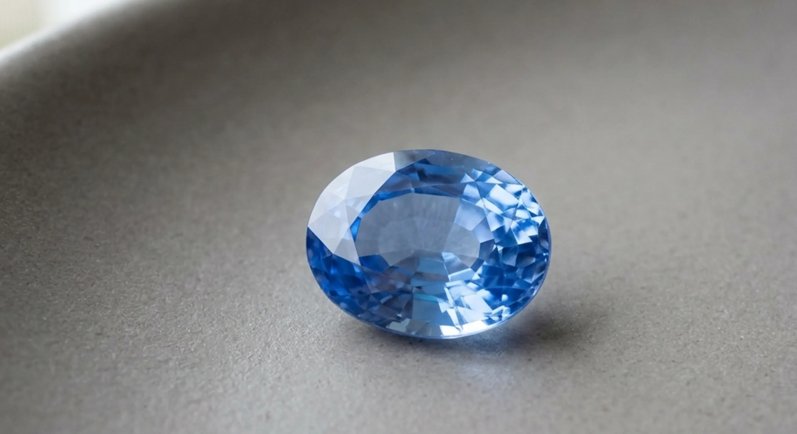 A single faceted cornflower-blue Ceylon sapphire showing bright, transparent colour on a neutral grey surface