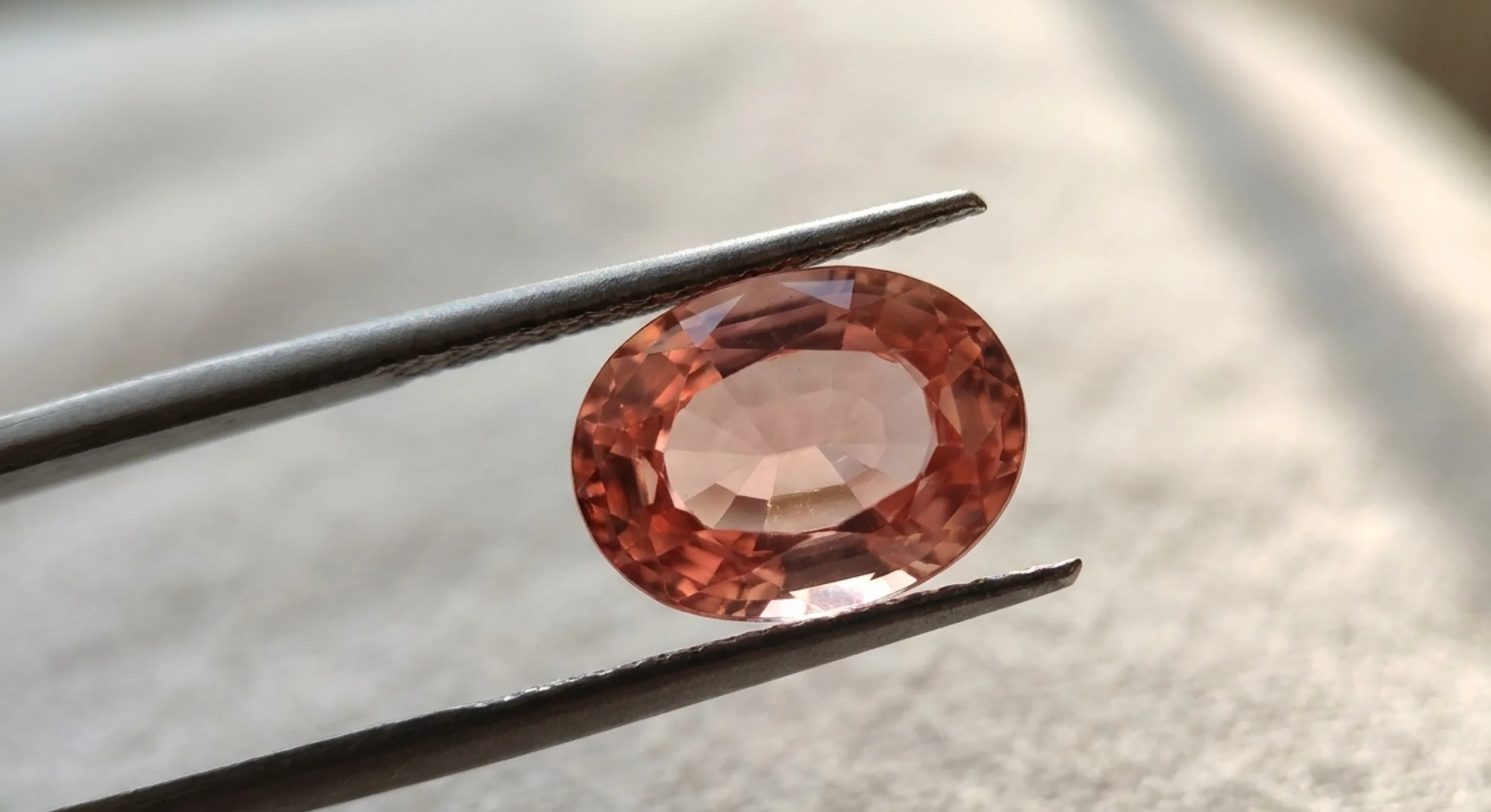 Loose oval peach sapphire held in tweezers showing an even warm pink-orange blush