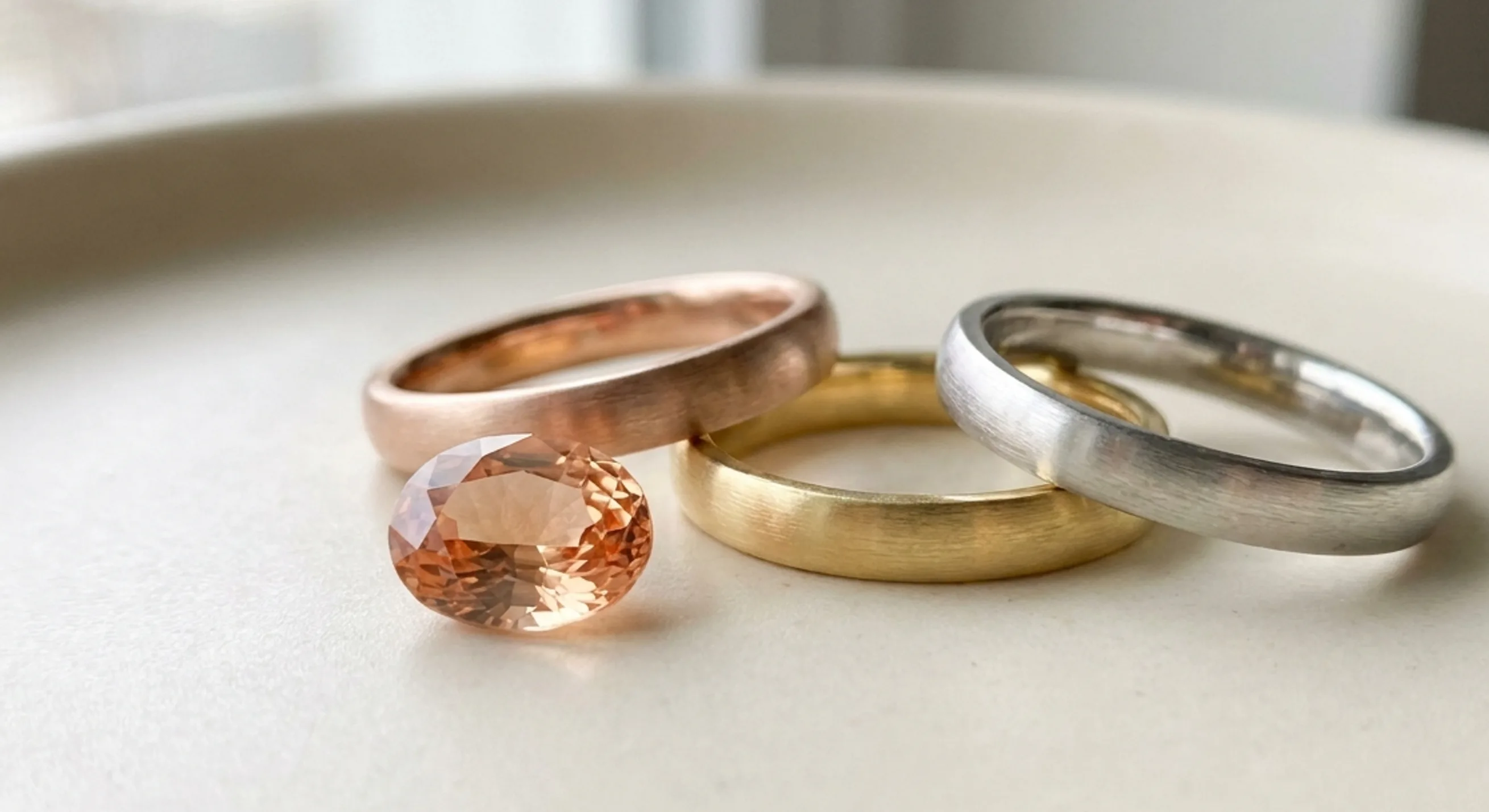 Loose peach sapphire beside rose gold, yellow gold, and platinum ring bands on a cream surface