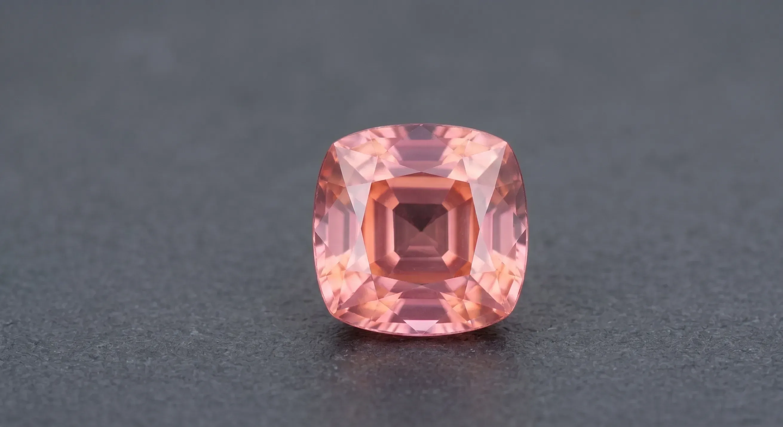 Cushion-cut faceted padparadscha sapphire in warm peach and pink tones on a dark textured surface under soft studio lighting.