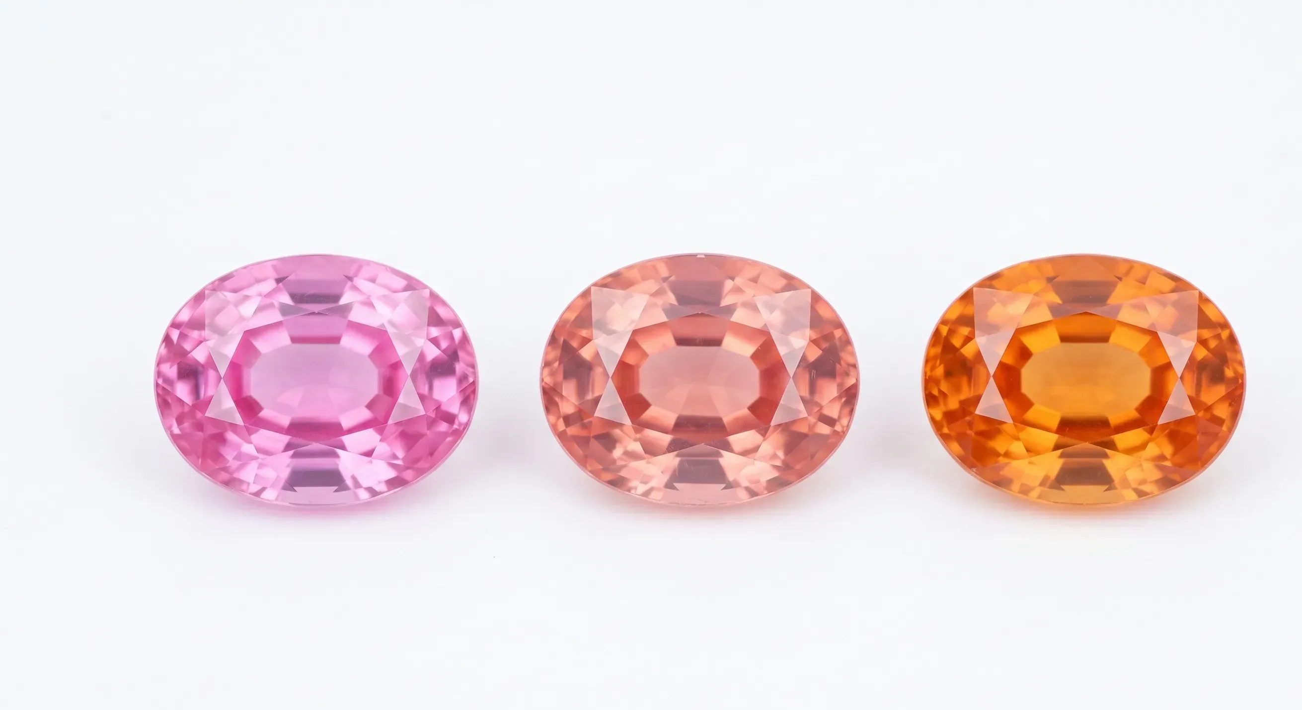 Three oval-cut faceted sapphires in a row on white: bright pink, peachy padparadscha, and deep orange.