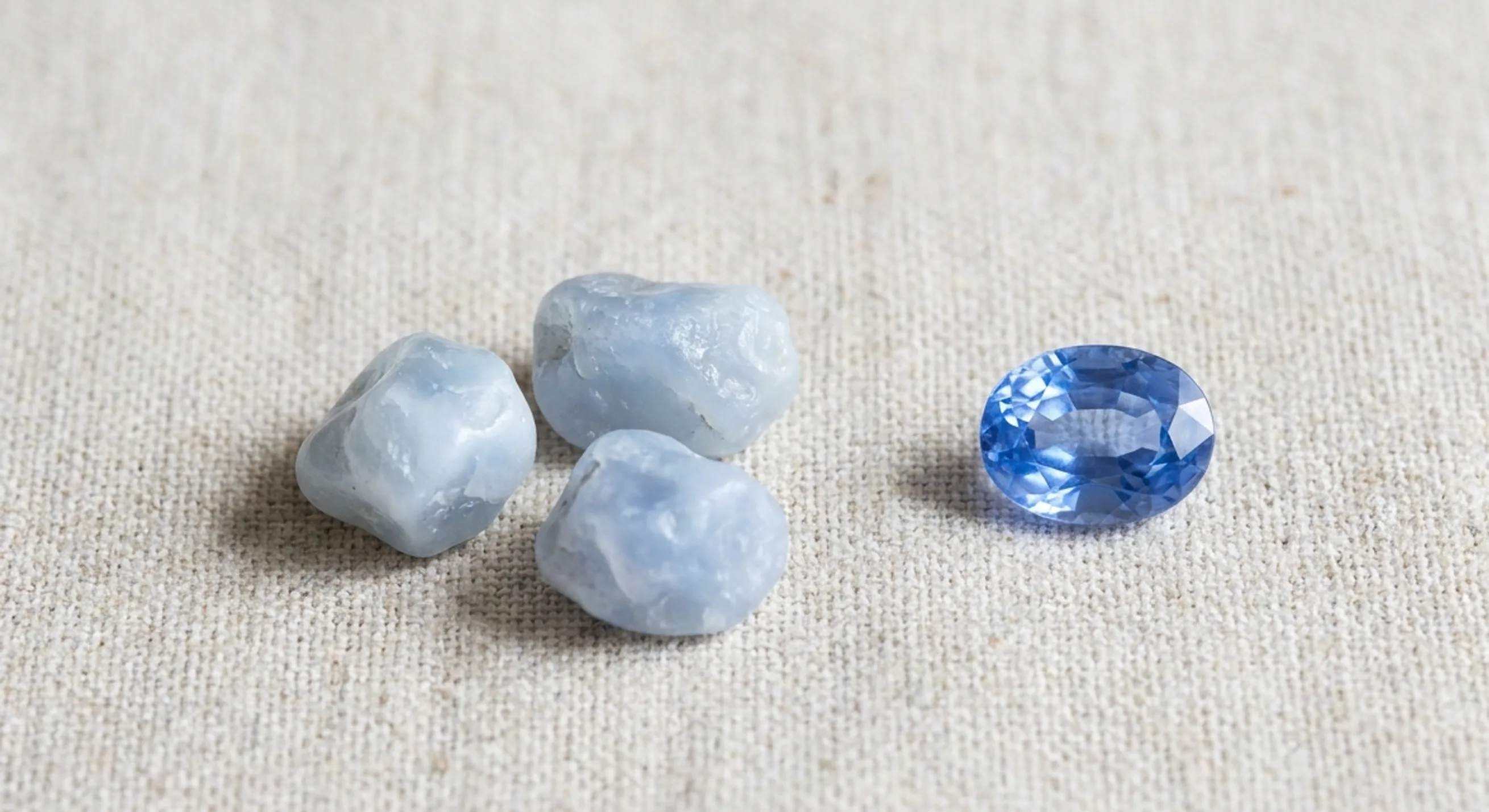 Milky pale geuda sapphire rough next to a faceted cornflower blue heated Ceylon sapphire on oatmeal linen