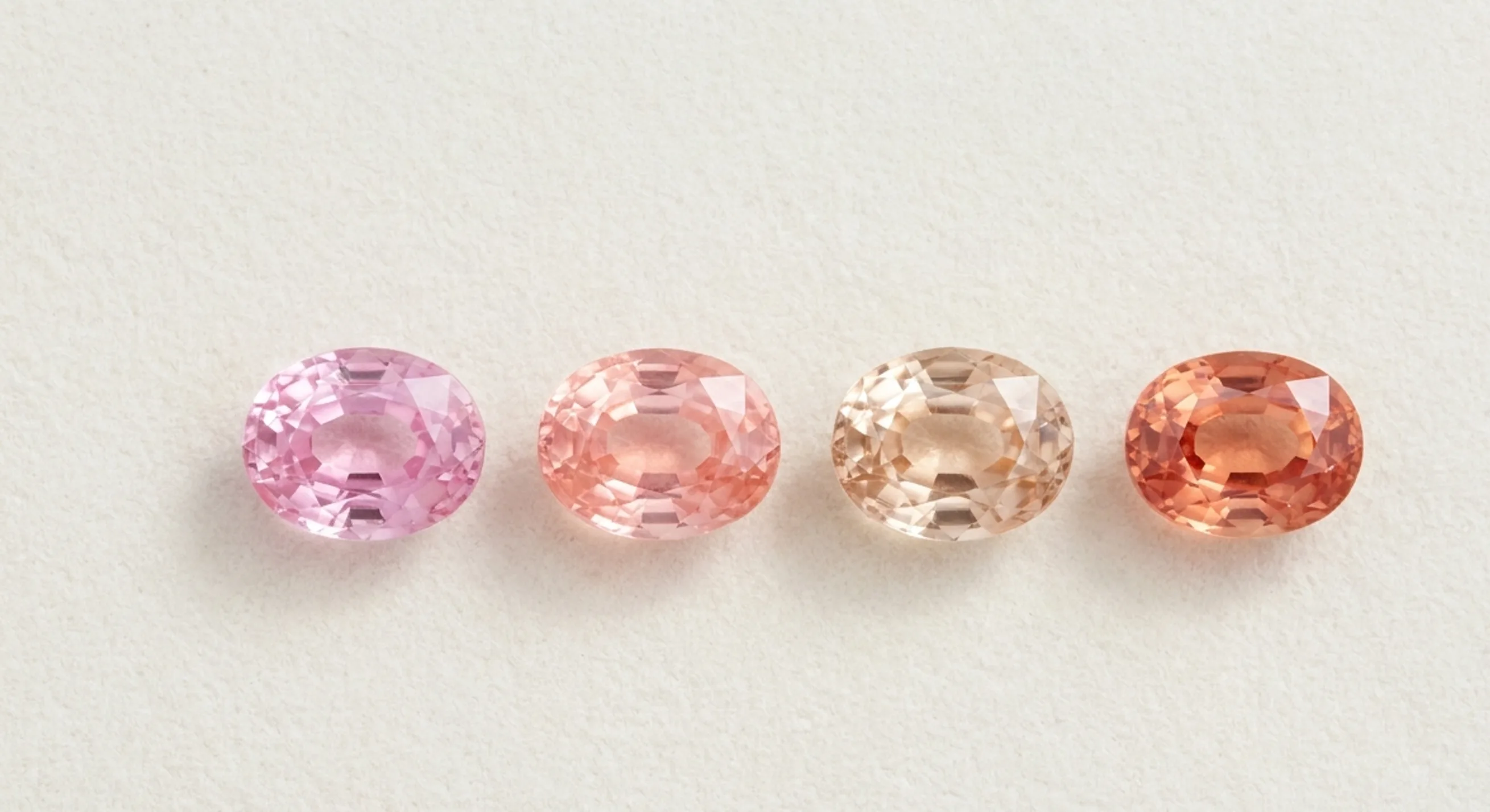 Pink, padparadscha, peach and salmon-orange sapphires in a row on pale linen, showing where padparadscha colour begins and ends