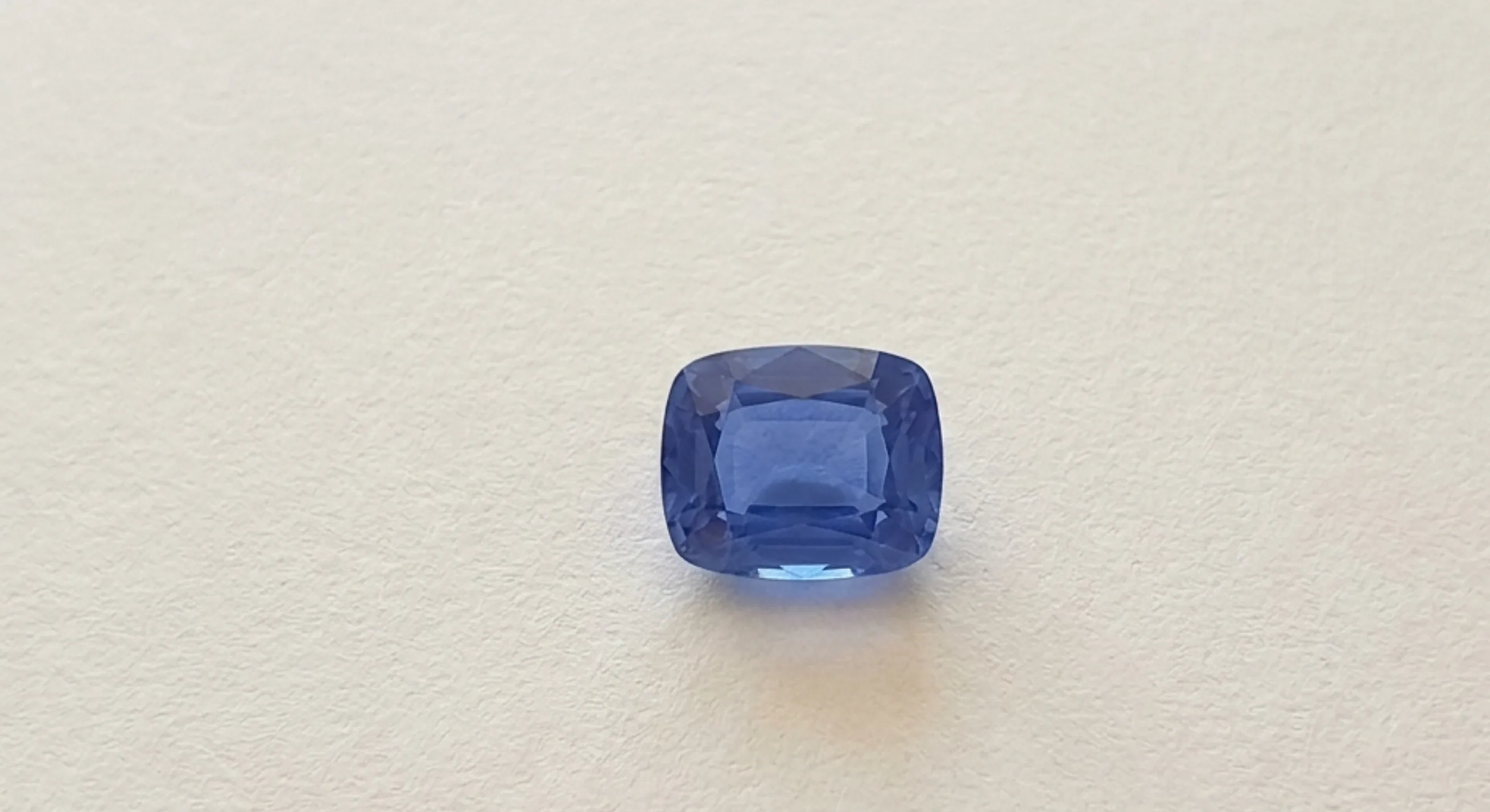 Cushion-cut blue sapphire lying face-up on white paper for a window test, with even colour across its face