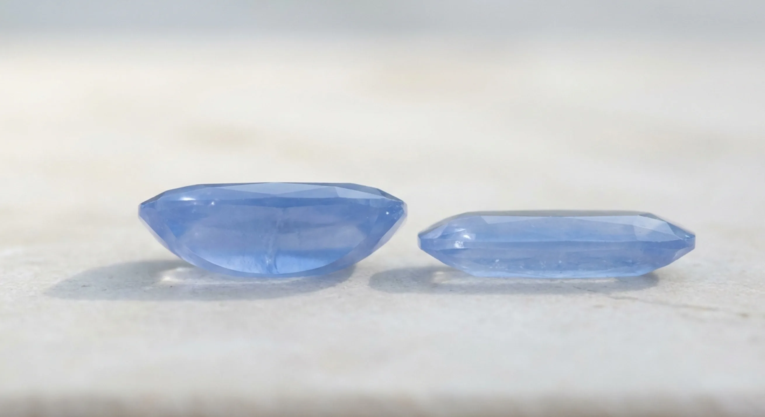 Profile view comparing a deep-pavilion cushion sapphire with a shallower light blue sapphire on pale stone