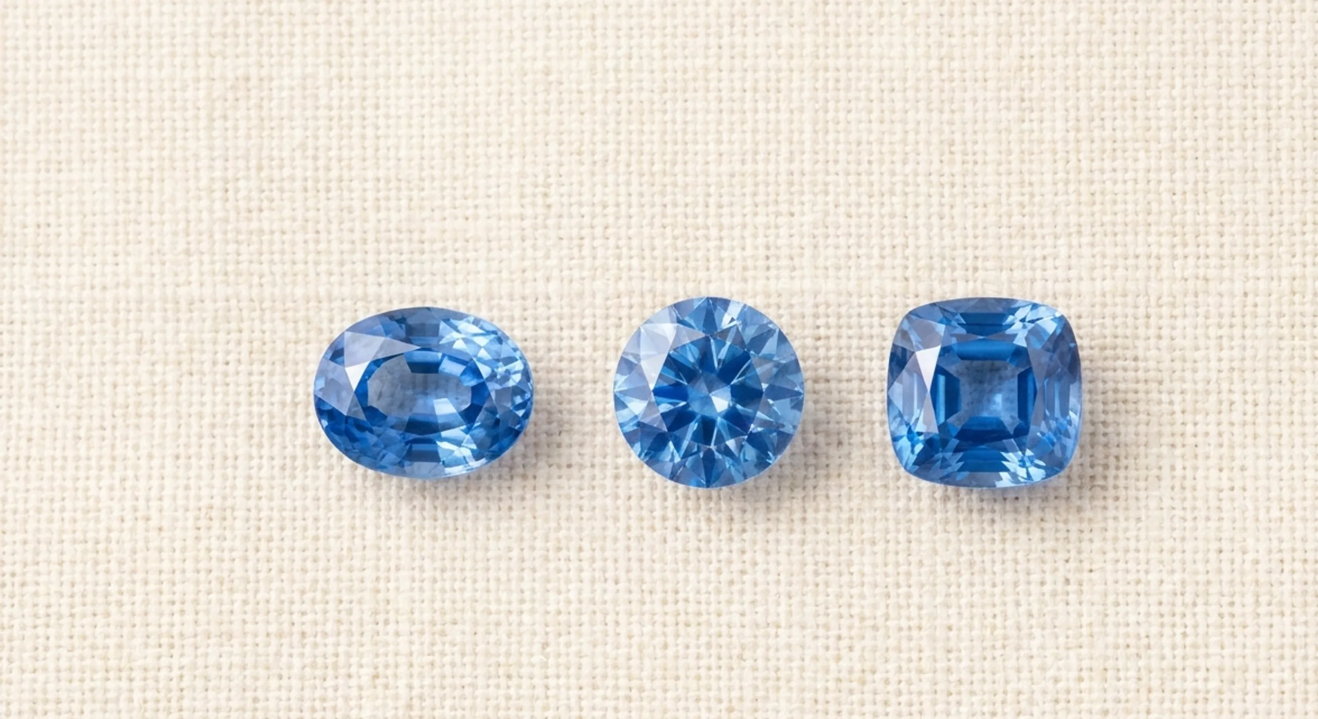 Oval, round and cushion-cut blue sapphires laid in a row on cream linen for shape comparison