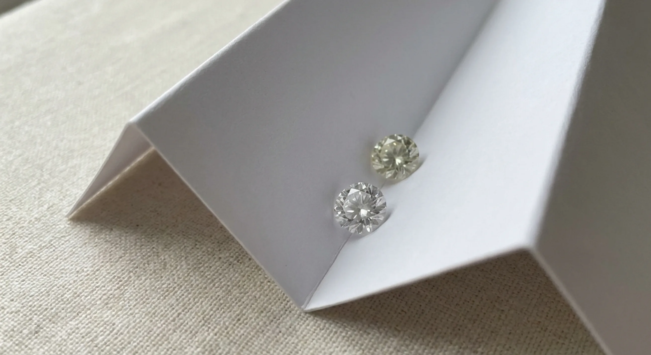 Two colorless gemstones table-down in a folded white card, one showing a faint warm tint