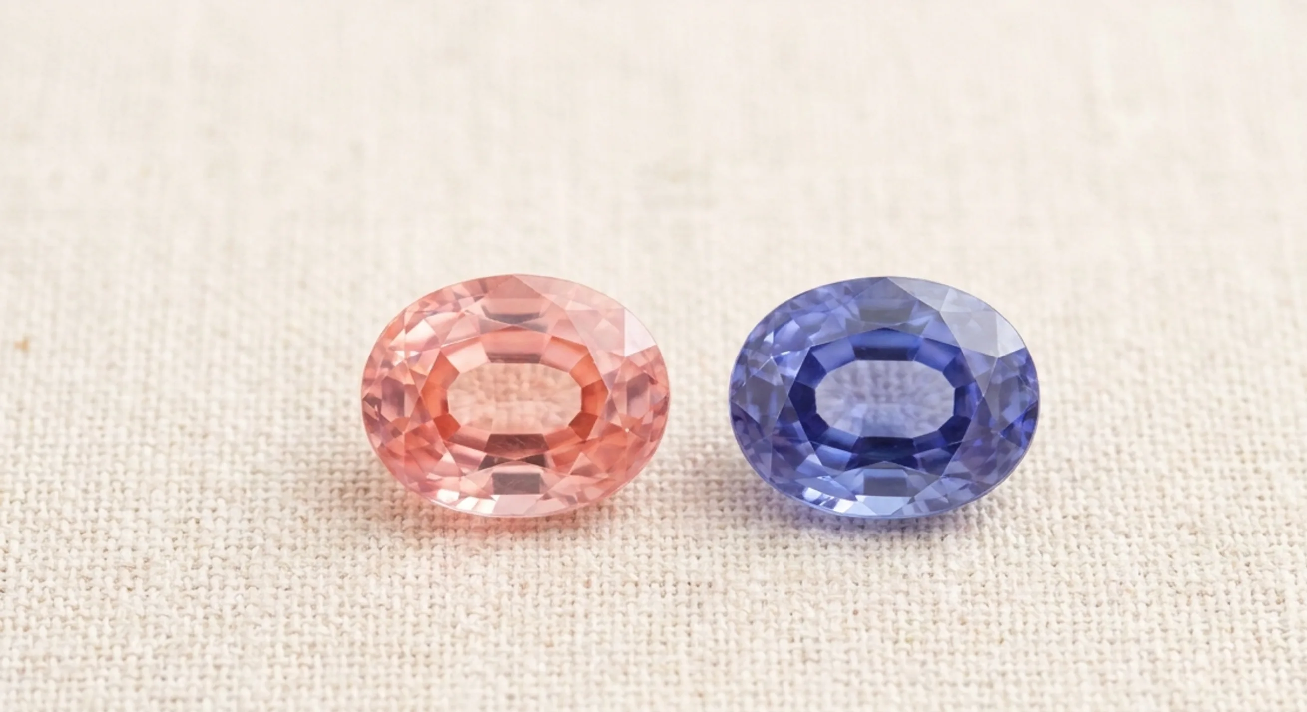 Salmon padparadscha sapphire next to a blue-violet colour-change sapphire on a pale linen tray in daylight