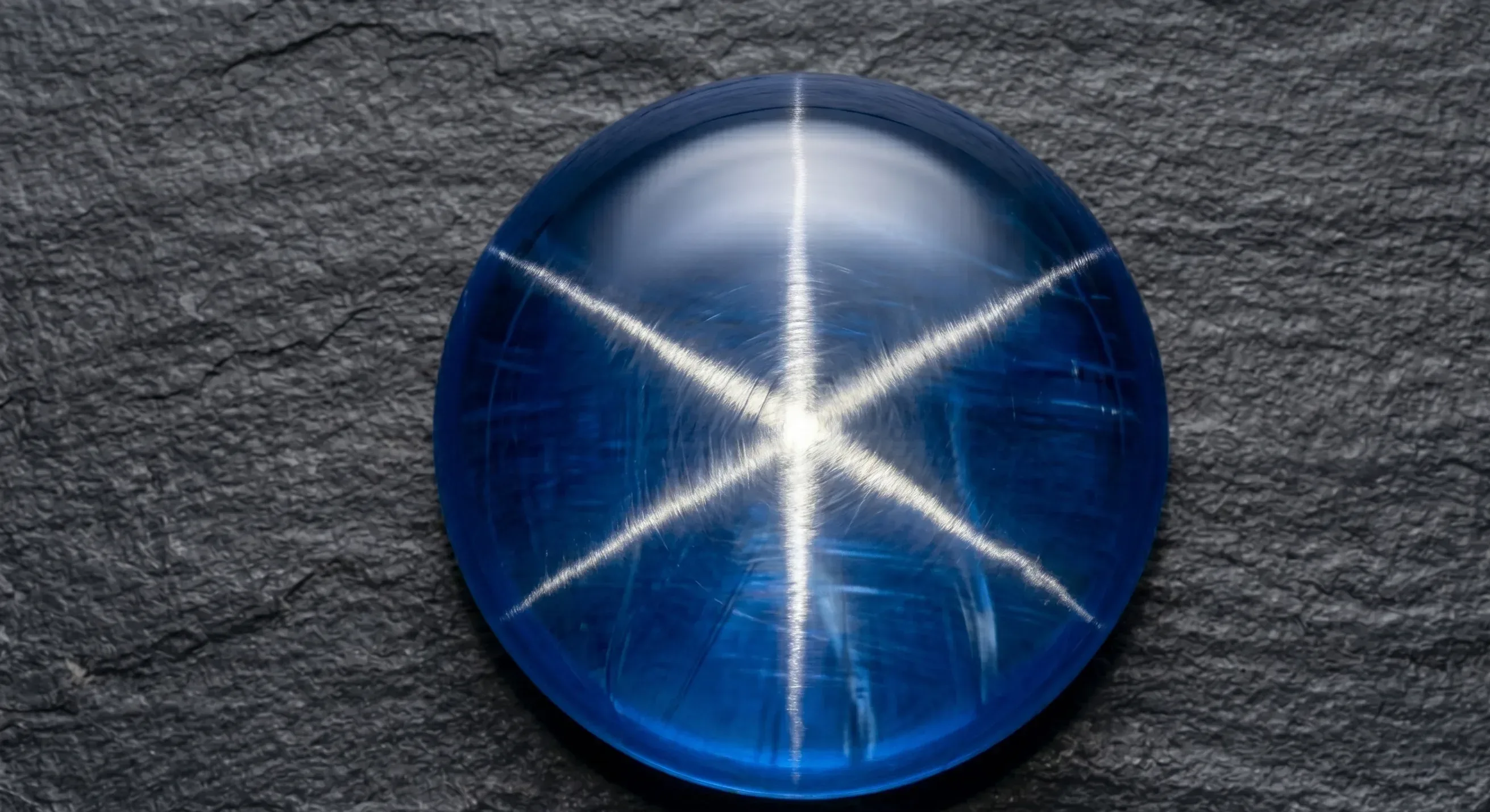 Macro overhead shot of a six-rayed cabochon star sapphire on black velvet, the three intersecting silver-white rays sharply defined under a single directional light.