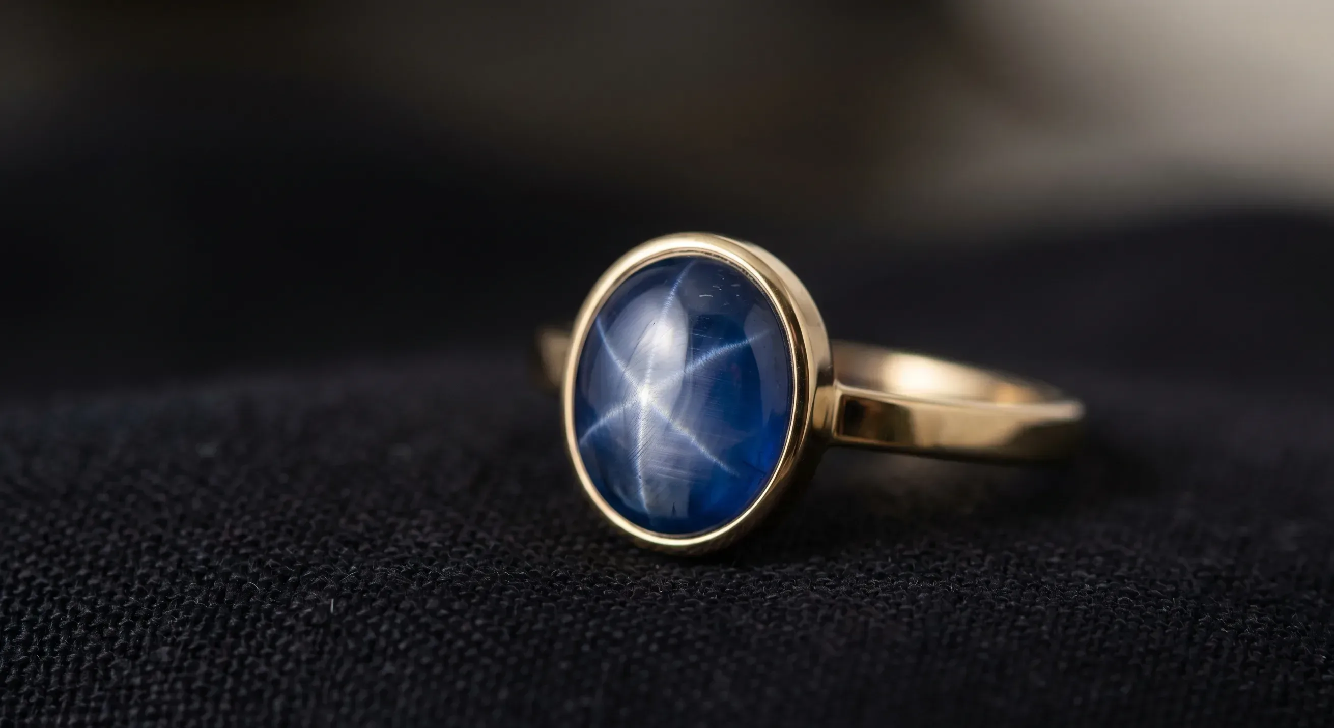 A cushion-style star sapphire cabochon set in a low-profile bezel ring on a black velvet surface, the star visible as the stone is tilted slightly into the light.