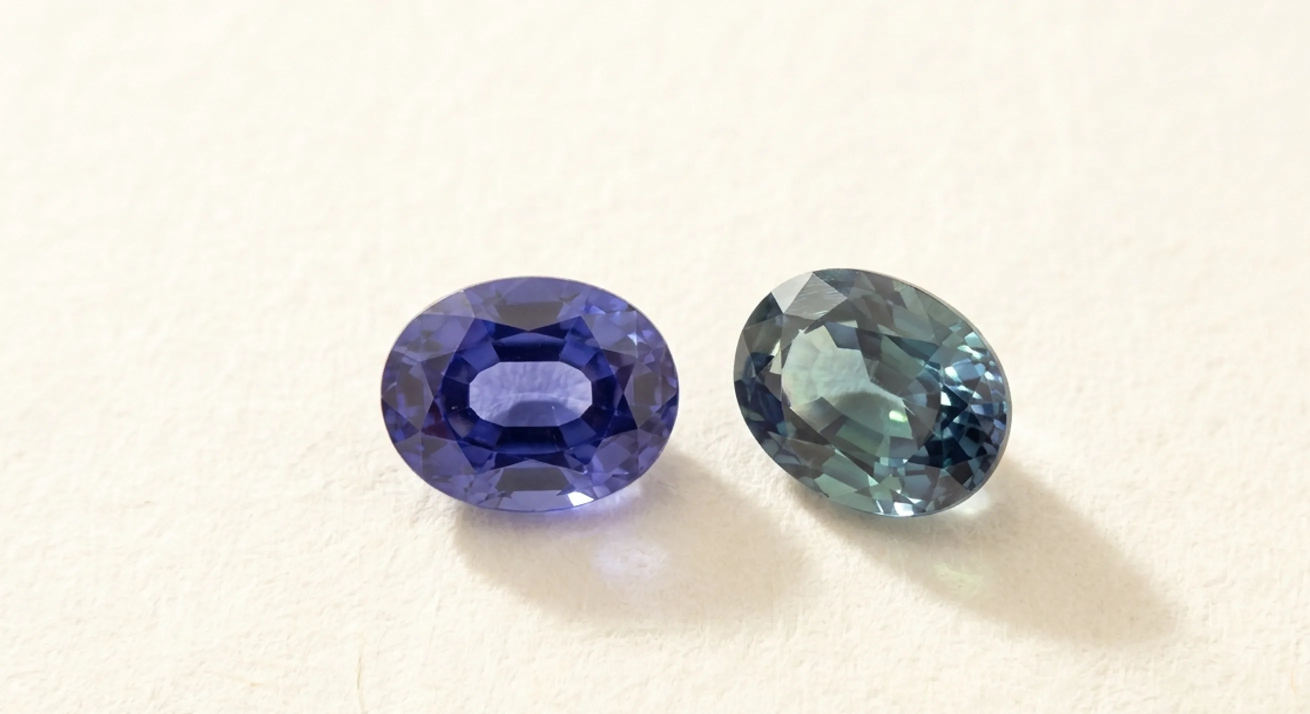 Two loose oval sapphires on white paper, one violetish blue face-up and one showing greenish-blue flashes