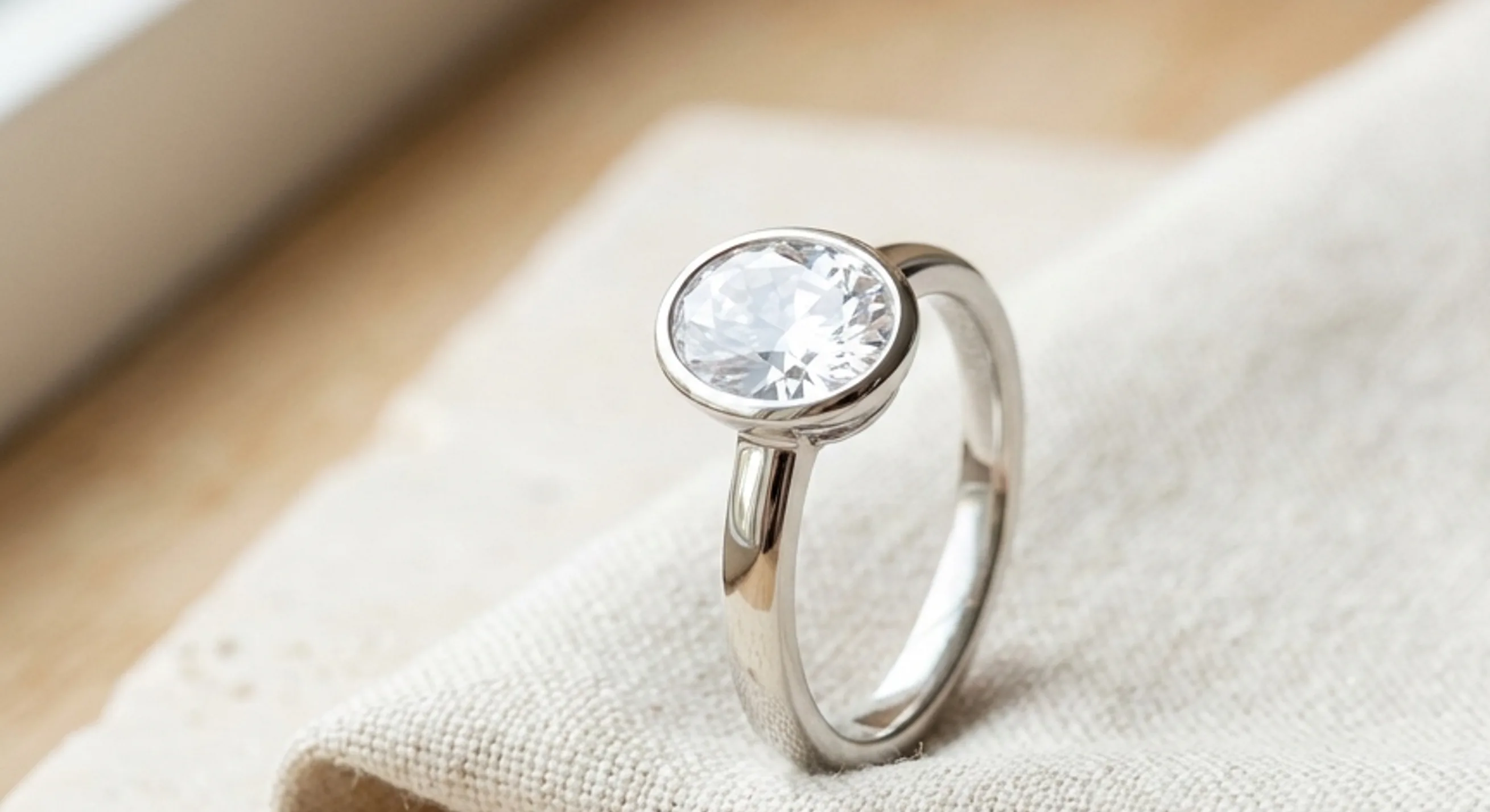 A white sapphire in a platinum bezel solitaire ring resting on bone-colored linen