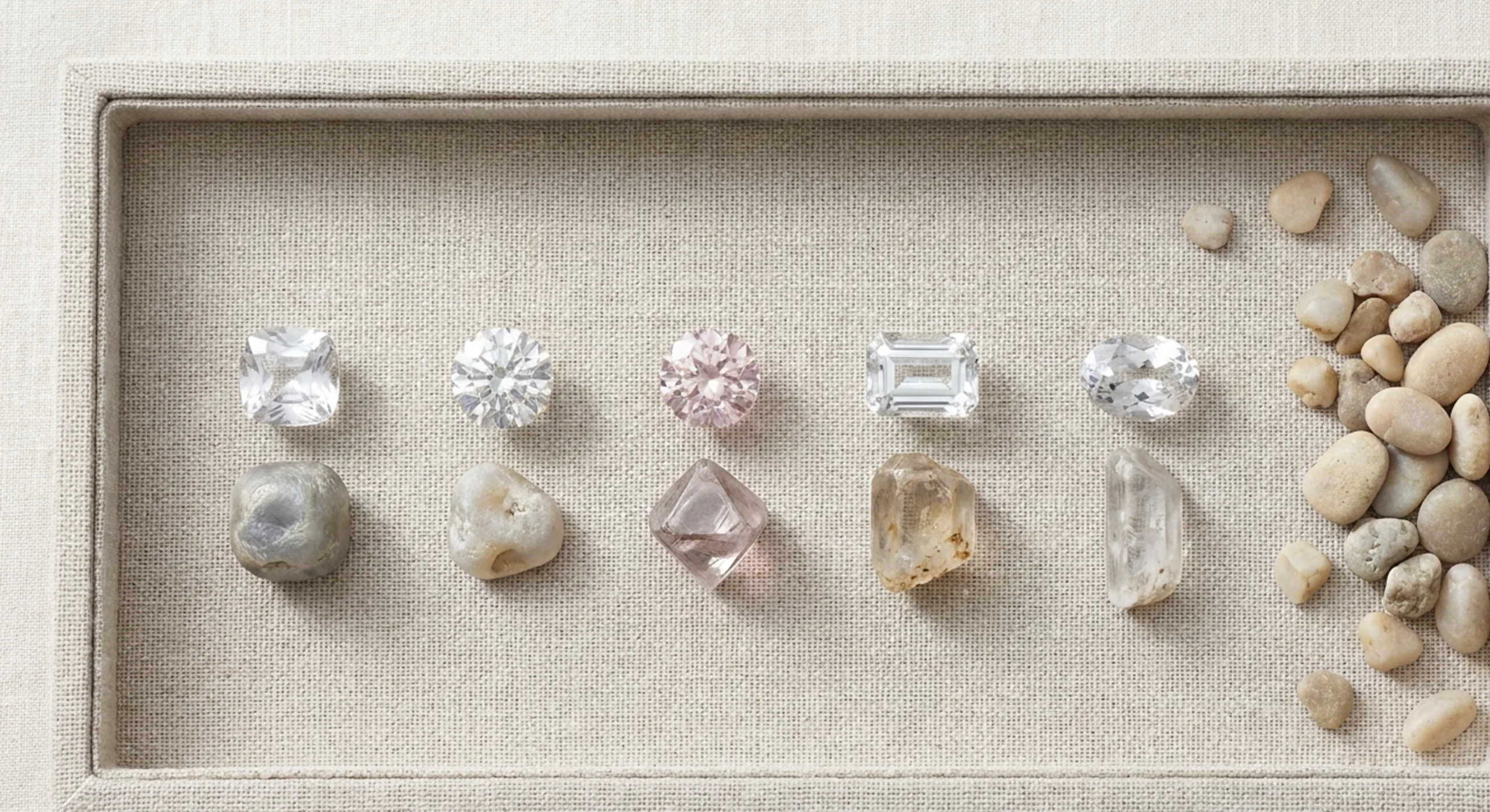Five cut Sri Lankan white gems beside their rough crystals and pale river gravel on linen